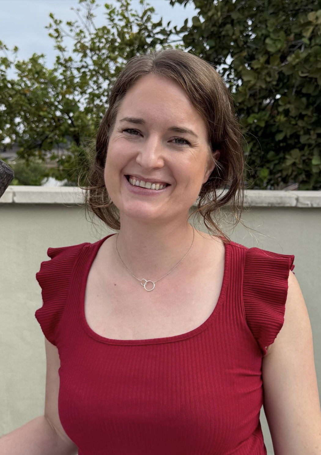 A woman with long brown hair, smiling and wearing a red top with ruffled sleeves, stands in front of a green backdrop with trees.